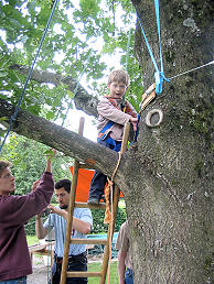 Foto: Ein kleiner Junge an einer Sicherungsleine klettert unter der Aufsicht eines Betreuers auf einen Baum